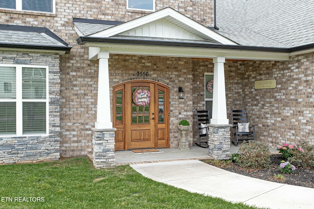 view of exterior entry with roof with shingles, a porch, board and batten siding, and brick siding