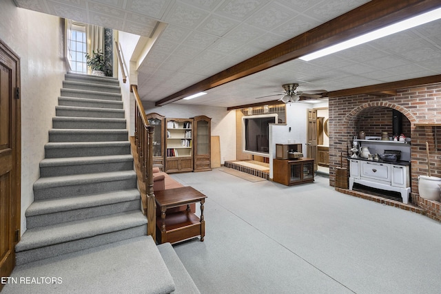 carpeted living area featuring brick wall, stairway, beam ceiling, a wood stove, and a ceiling fan