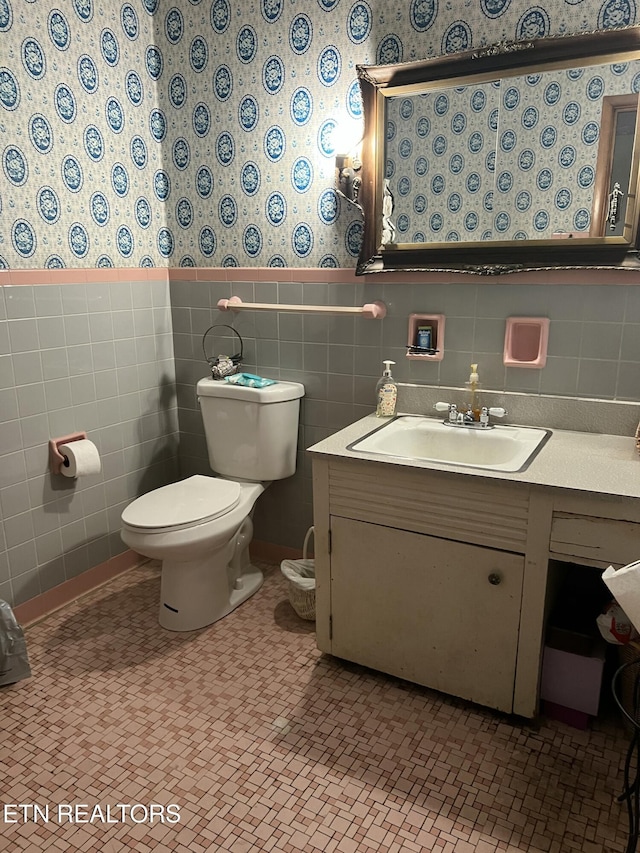 bathroom featuring wallpapered walls, toilet, a wainscoted wall, vanity, and tile walls