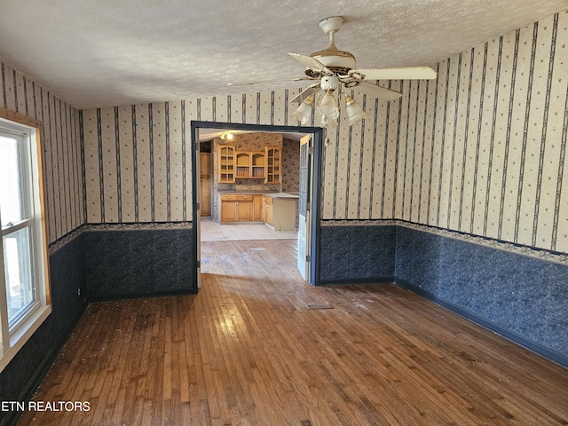 empty room with a textured ceiling, ceiling fan, hardwood / wood-style floors, and wallpapered walls