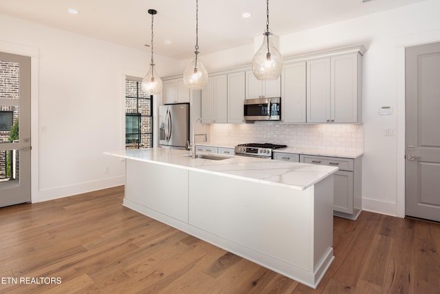 kitchen with wood finished floors, a sink, appliances with stainless steel finishes, tasteful backsplash, and a center island with sink