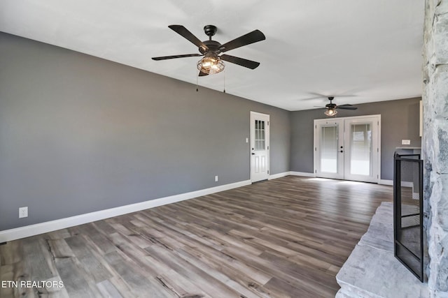 unfurnished living room with ceiling fan, a stone fireplace, wood finished floors, baseboards, and french doors