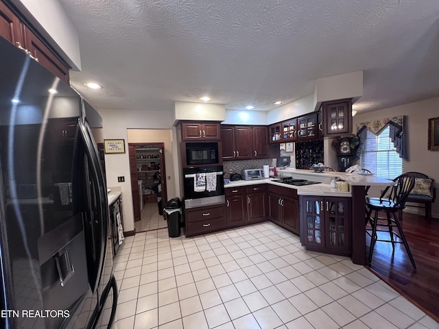 kitchen featuring a peninsula, dark brown cabinets, black appliances, tasteful backsplash, and glass insert cabinets