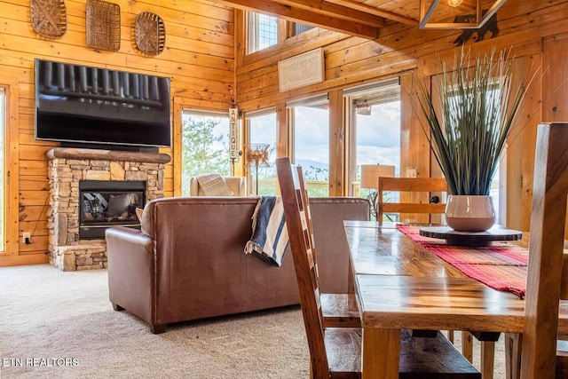 carpeted dining room with plenty of natural light, wooden walls, beam ceiling, and a stone fireplace