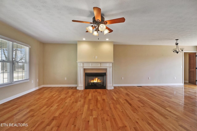 unfurnished living room with baseboards, a warm lit fireplace, ceiling fan with notable chandelier, and light wood finished floors