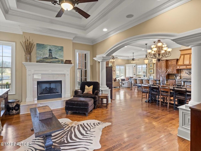 living area featuring a raised ceiling, light wood finished floors, ceiling fan with notable chandelier, and ornate columns