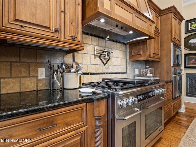 kitchen featuring brown cabinetry, custom exhaust hood, stainless steel appliances, decorative backsplash, and light wood-type flooring