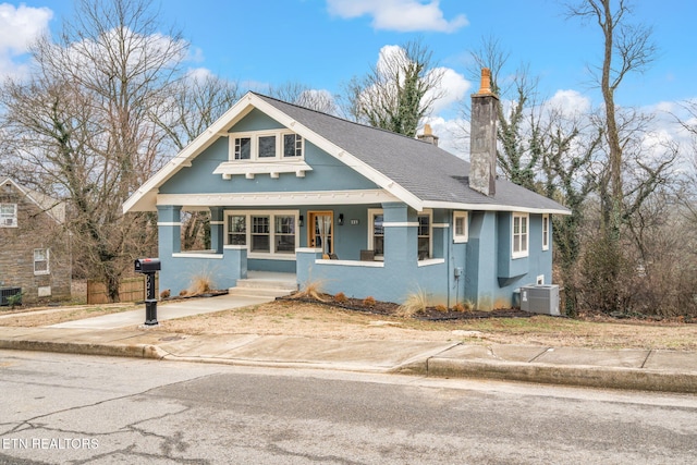 view of front facade with central AC unit, roof with shingles, a porch, a chimney, and stucco siding