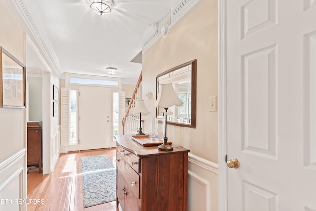 foyer entrance featuring crown molding, a wainscoted wall, stairs, light wood-style floors, and a decorative wall