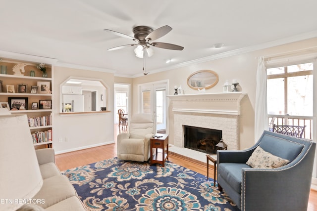 living room featuring wood finished floors, baseboards, a ceiling fan, a fireplace, and crown molding