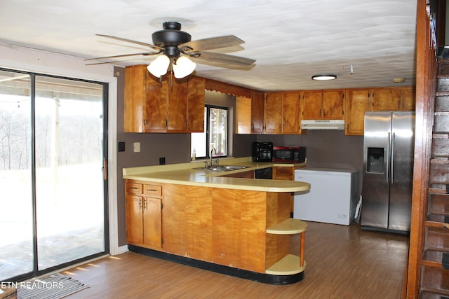 kitchen featuring brown cabinetry, a peninsula, a sink, stainless steel refrigerator with ice dispenser, and under cabinet range hood