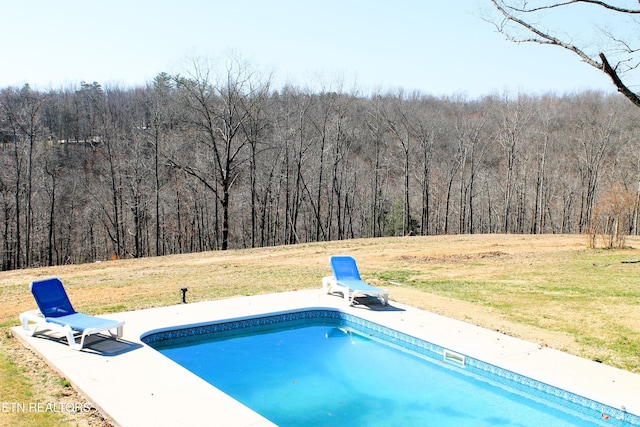 outdoor pool with a patio area, a lawn, and a wooded view