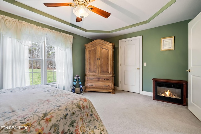 bedroom with baseboards, ceiling fan, light colored carpet, a tray ceiling, and a glass covered fireplace