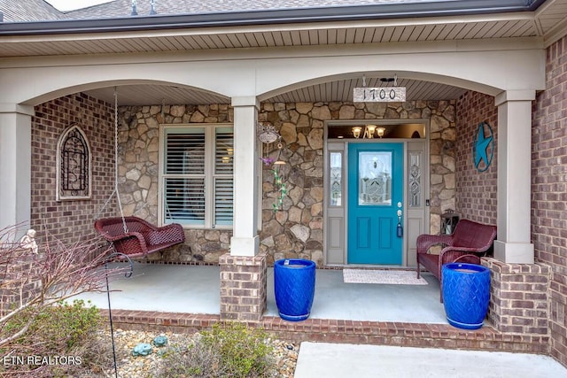 property entrance with covered porch, stone siding, and roof with shingles