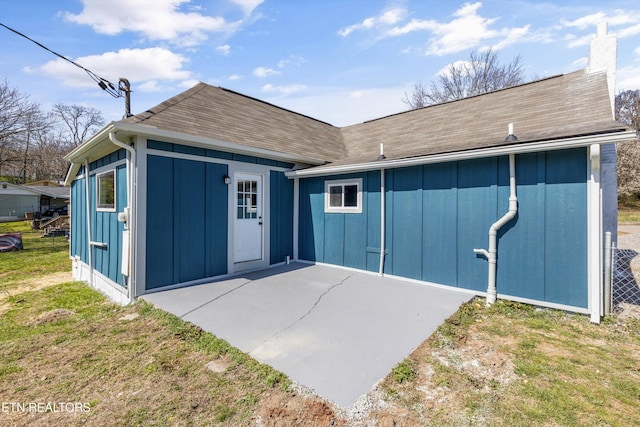 back of house with a lawn, board and batten siding, a shingled roof, a chimney, and a patio area