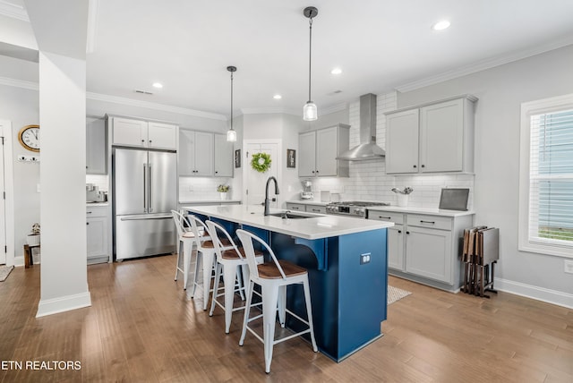 kitchen with a kitchen bar, a center island with sink, a sink, appliances with stainless steel finishes, and wall chimney range hood