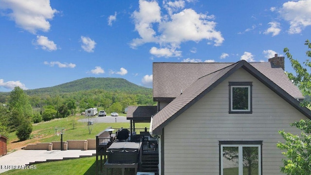 view of property exterior featuring a yard, a deck with mountain view, a chimney, and a shingled roof