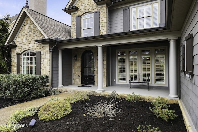 view of exterior entry with stone siding, a porch, and roof with shingles