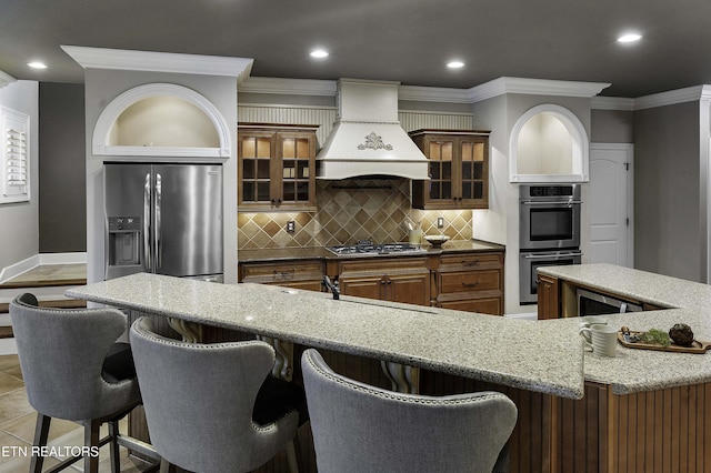 kitchen featuring light stone countertops, custom exhaust hood, stainless steel appliances, crown molding, and brown cabinets