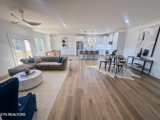 living room with baseboards, light wood finished floors, recessed lighting, crown molding, and ceiling fan with notable chandelier