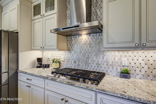 kitchen featuring light stone counters, stainless steel appliances, glass insert cabinets, wall chimney exhaust hood, and tasteful backsplash