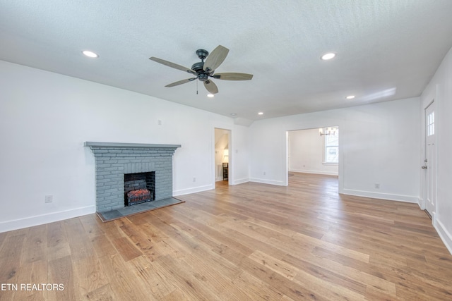 unfurnished living room featuring ceiling fan with notable chandelier, a brick fireplace, light wood-style floors, and a textured ceiling