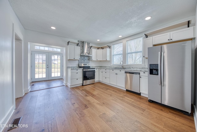 kitchen featuring light wood finished floors, a sink, stainless steel appliances, french doors, and wall chimney exhaust hood