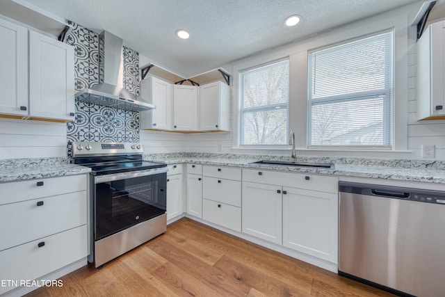 kitchen with a sink, light wood-style floors, appliances with stainless steel finishes, white cabinetry, and wall chimney range hood