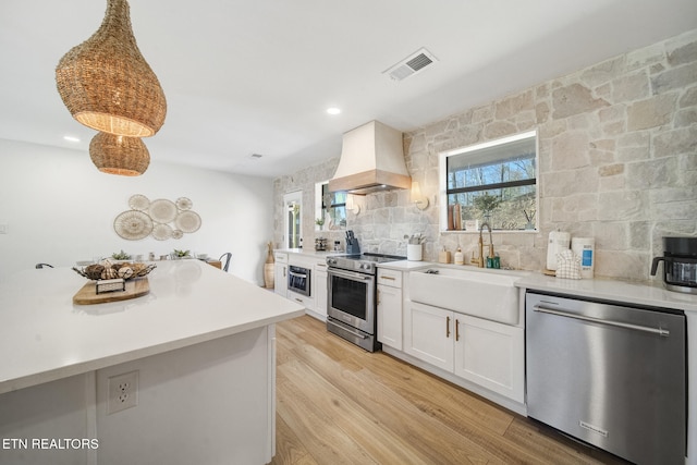 kitchen featuring visible vents, custom exhaust hood, stainless steel appliances, white cabinets, and light wood-style floors