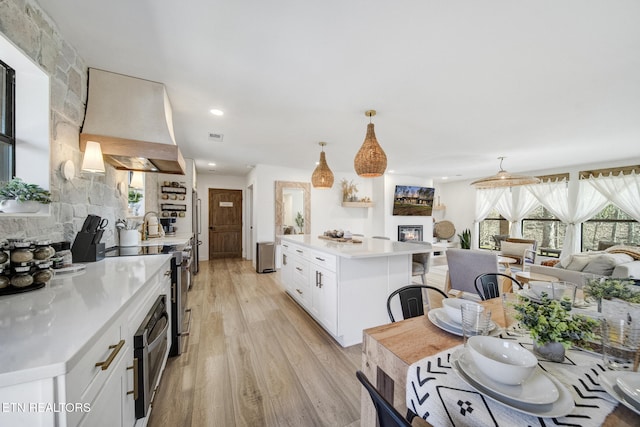 kitchen with premium range hood, open floor plan, a glass covered fireplace, white cabinets, and light wood finished floors