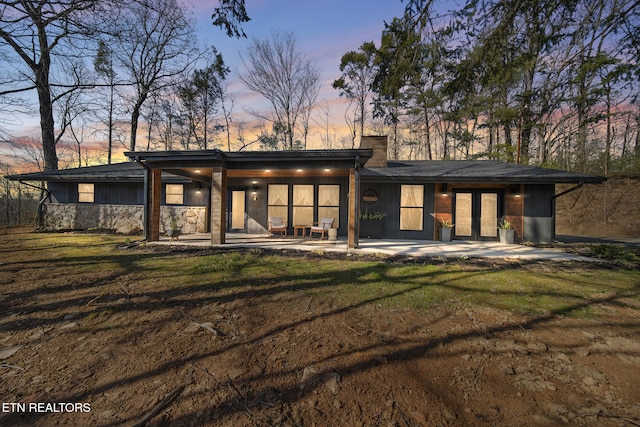 back of property at dusk featuring french doors, stone siding, a lawn, and a chimney