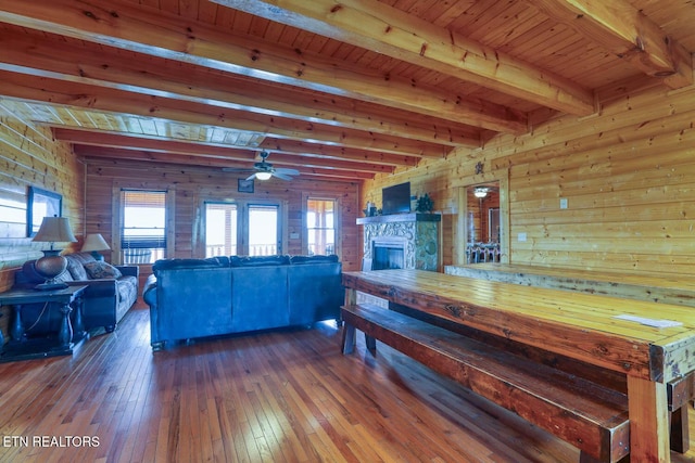 unfurnished dining area with beamed ceiling, wood walls, a stone fireplace, hardwood / wood-style flooring, and a ceiling fan