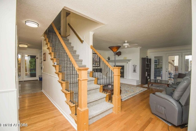 stairs with wood finished floors, a wainscoted wall, a fireplace with raised hearth, ceiling fan, and a textured ceiling