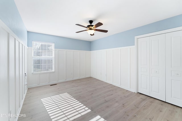 unfurnished bedroom featuring visible vents, ceiling fan, a wainscoted wall, light wood-style flooring, and a decorative wall