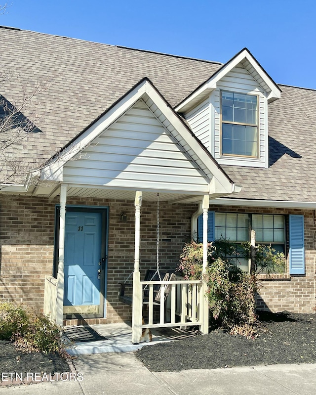 property entrance featuring brick siding, covered porch, and a shingled roof