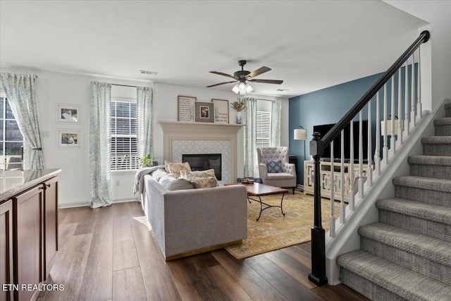 living area with stairway, a ceiling fan, visible vents, a tile fireplace, and dark wood-type flooring