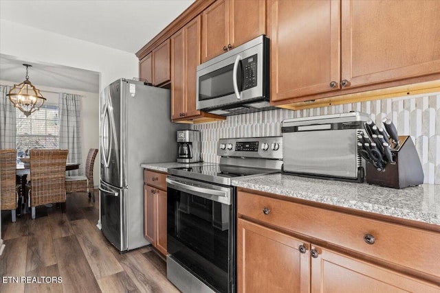 kitchen featuring brown cabinetry, decorative backsplash, dark wood-type flooring, appliances with stainless steel finishes, and a notable chandelier