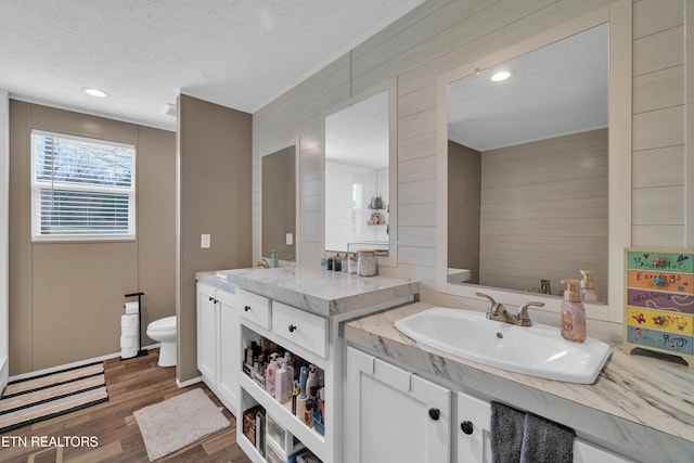 full bathroom featuring toilet, two vanities, wood finished floors, a textured ceiling, and a sink