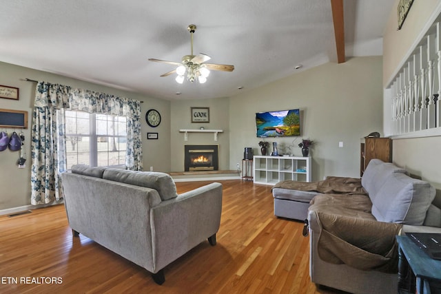 living room with light wood-type flooring, visible vents, a glass covered fireplace, ceiling fan, and vaulted ceiling with beams