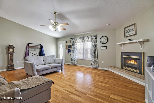 living area featuring light wood-type flooring, a glass covered fireplace, baseboards, ceiling fan, and vaulted ceiling