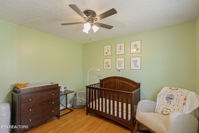 bedroom featuring baseboards, a nursery area, wood finished floors, a textured ceiling, and a ceiling fan