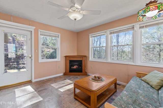 living area featuring a glass covered fireplace, baseboards, ceiling fan, and stone finish floor