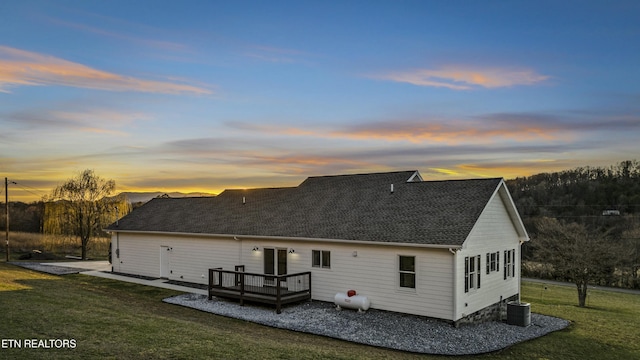 back of house at dusk featuring central air condition unit, a deck, a yard, roof with shingles, and a patio area