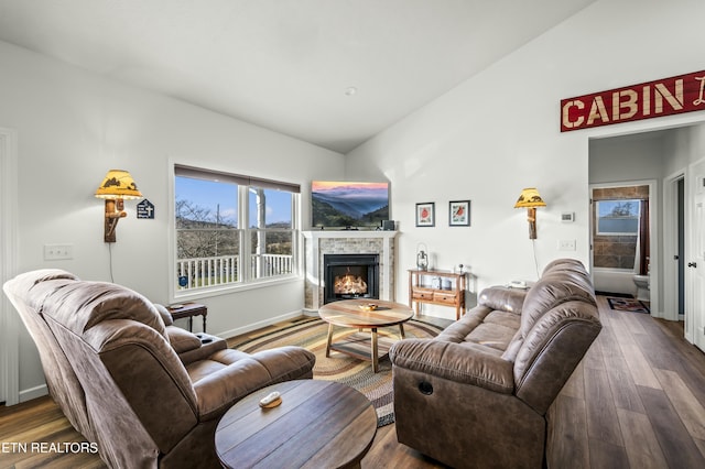 living area with a warm lit fireplace, wood finished floors, baseboards, and vaulted ceiling