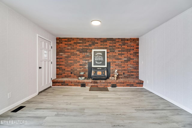 unfurnished living room featuring wood finished floors, visible vents, brick wall, baseboards, and a wood stove