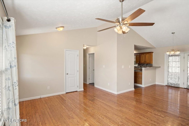 unfurnished living room with ceiling fan with notable chandelier, high vaulted ceiling, light wood-type flooring, and baseboards