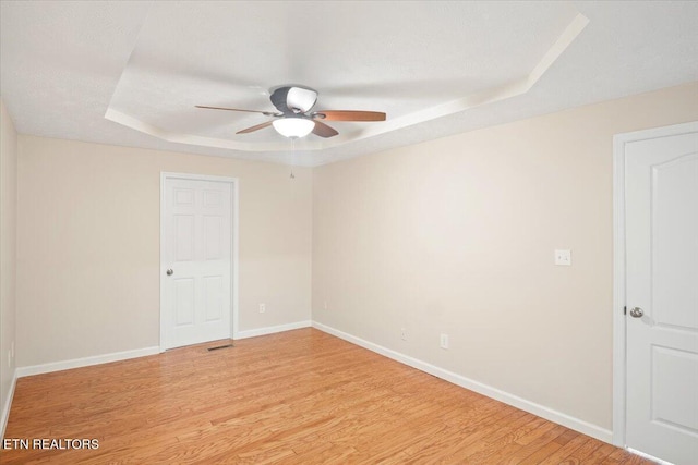 empty room with light wood-type flooring, a raised ceiling, baseboards, and ceiling fan