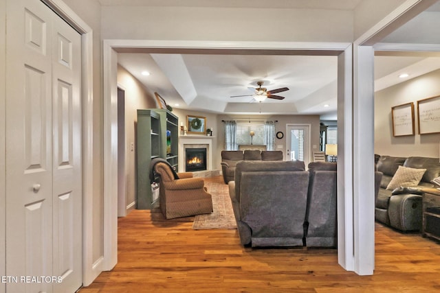 living area featuring a glass covered fireplace, a tray ceiling, wood finished floors, and ceiling fan