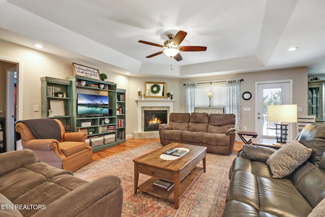 living room featuring a ceiling fan, a tray ceiling, recessed lighting, a lit fireplace, and light wood finished floors