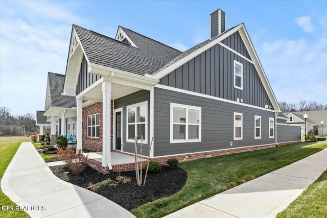 view of side of property with roof with shingles, a porch, a chimney, a lawn, and board and batten siding